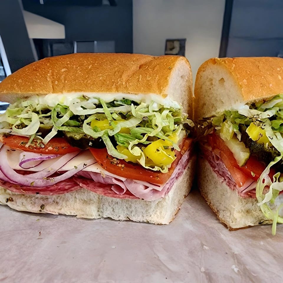 A large Yellow Mustard sandwich, cut in half, filled with meats, cheese, lettuce, tomatoes, and onions.