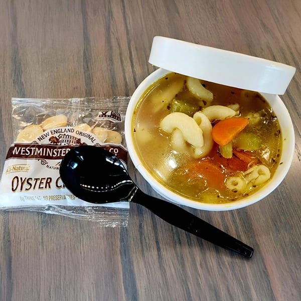 A bowl of Yellow Mustard's chicken noodle soup with a side of oyster crackers and a spoon.