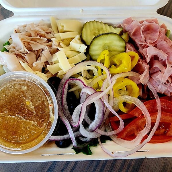 Deli salad with turkey, ham, cheese, pickles, onions, tomatoes, and dressing in a white container.